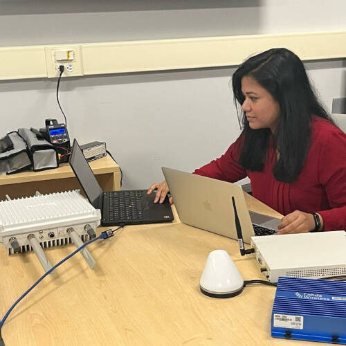 Dr. Naureen wearing a red top is seated at a desk working on two laptops. In front of her are various electronic and networking devices, including a white router-like unit with multiple antennas, a small white dome-shaped device.